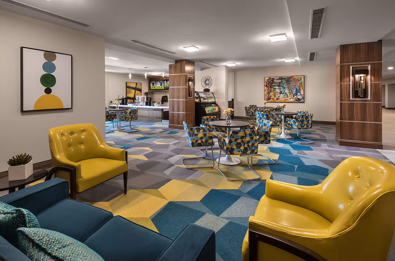 Bright senior living common area with geometric carpet, yellow and teal lounge chairs, round tables, and a small service counter in the background.
