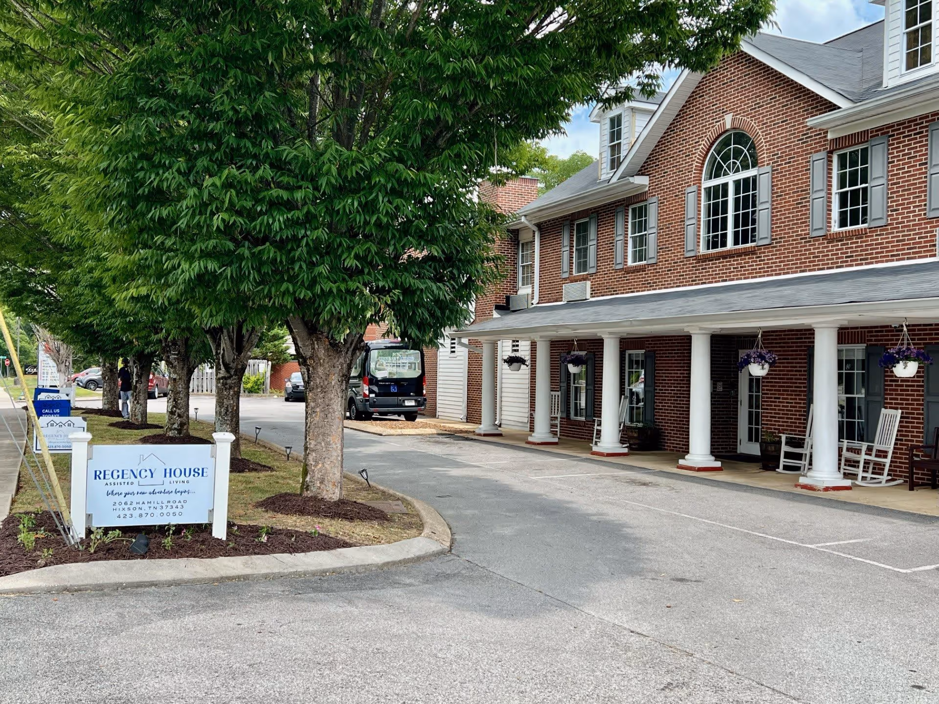 Exterior view of Regency House assisted living facility showing a brick building with white columns and hanging flower pots. There are several trees along the driveway and a sign with the facility's name and contact information.