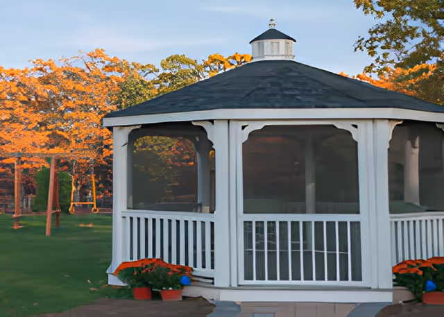 White screened gazebo on a lawn with autumn trees and potted flowers.