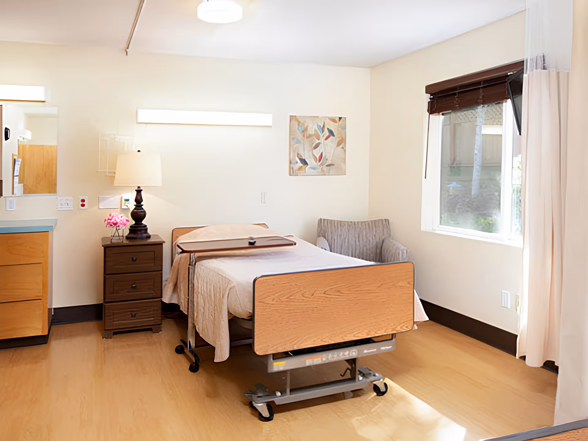 Well-lit senior facility patient bedroom with a hospital-style bed, nightstand and lamp, armchair and window.