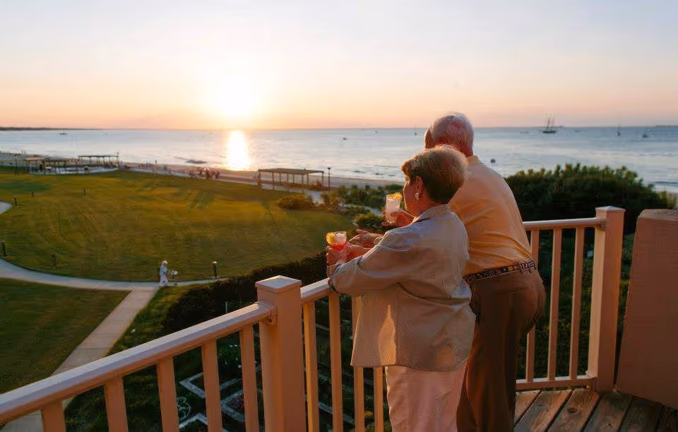 An elderly couple standing on a wooden balcony overlooking a grassy area and a body of water at sunset. They are holding drinks and enjoying the scenic view with boats visible on the water.