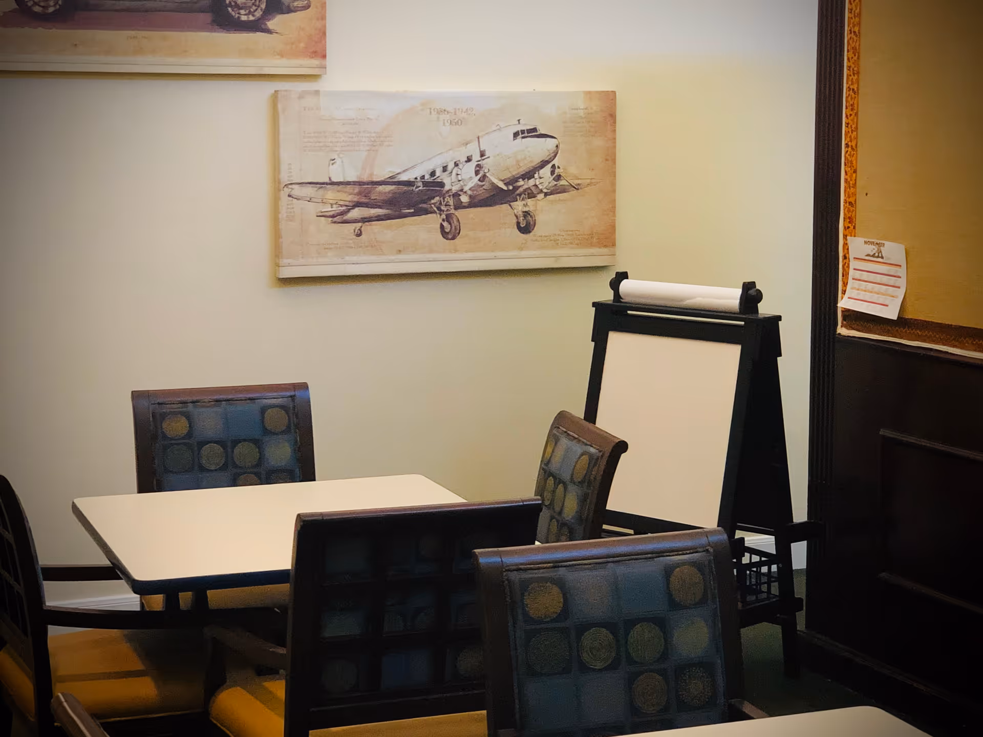 Small dining/activity room with a square table surrounded by patterned chairs, a flip-chart easel, and airplane wall art on the wall.