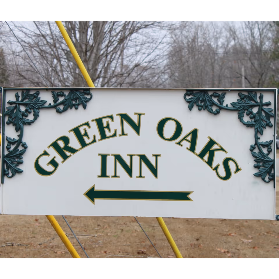 A white sign with green decorative leaves in the corners displaying the text 'GREEN OAKS INN' with a left-pointing arrow beneath the text. The background shows leafless trees and a grassy area.