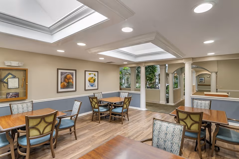 A well-lit dining area with wooden tables and cushioned chairs arranged neatly. The room features a light wood floor, beige and blue walls, framed fruit artwork on the wall, and large skylights in the ceiling. White columns and an indoor garden area are visible in the background.