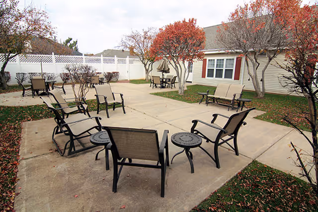 Outdoor patio area with several metal chairs and small round tables arranged on a concrete surface. There are trees with red and orange leaves, a white fence in the background, and a building with beige siding and white-framed windows.