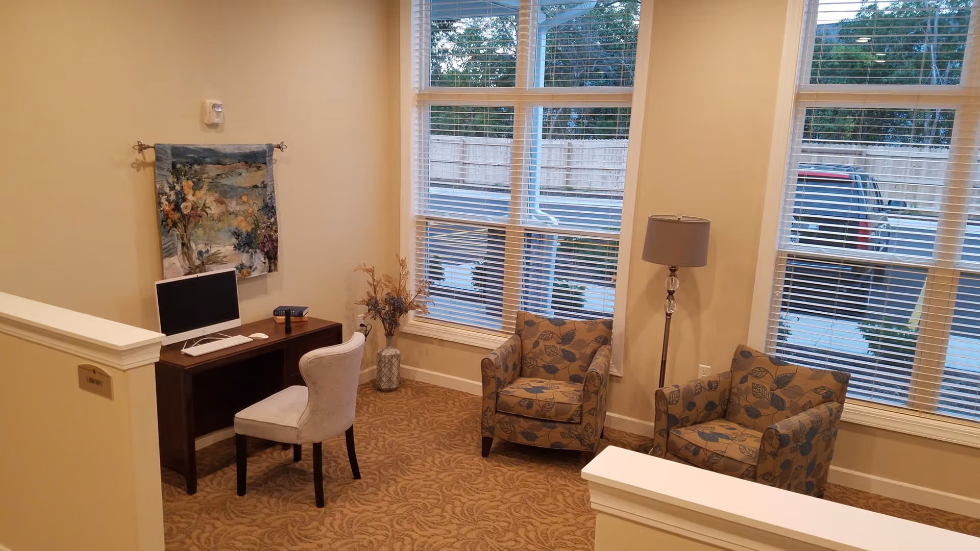 A cozy interior corner of a senior living facility with a small wooden desk holding a computer and a chair. Two patterned armchairs and a floor lamp are positioned near large windows with blinds, allowing natural light to enter. A decorative wall hanging and a vase with dried flowers add a homey touch.
