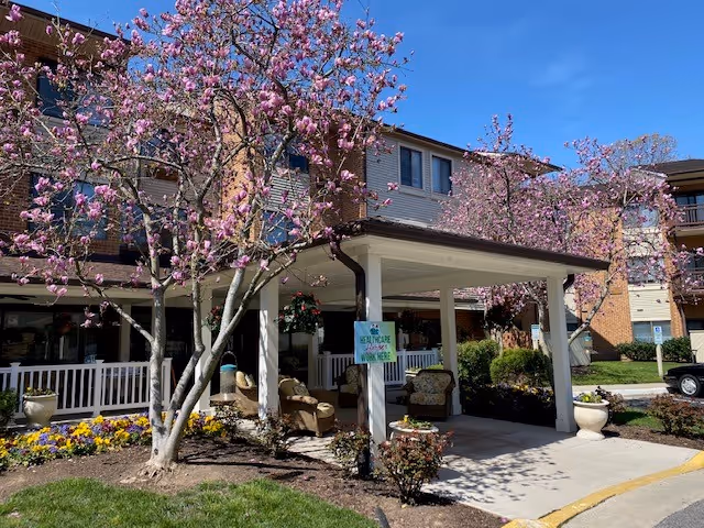 Exterior view of Potomac Place Assisted Living and Memory Care building with blooming pink flowering trees and a covered entrance area featuring outdoor seating and flower beds under a clear blue sky.