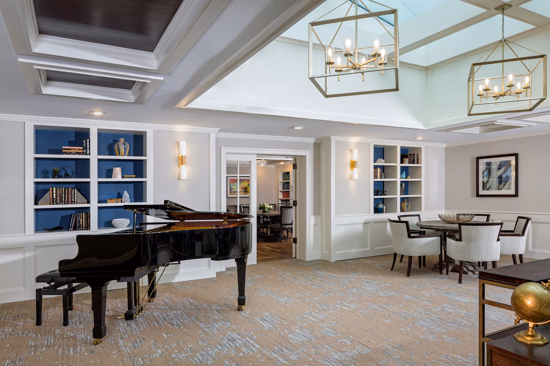 A spacious, well-lit common area featuring a black grand piano on the left, built-in white shelves with blue back panels holding decorative items and books, a round table with six upholstered chairs on the right, and two modern rectangular chandeliers hanging from the ceiling.