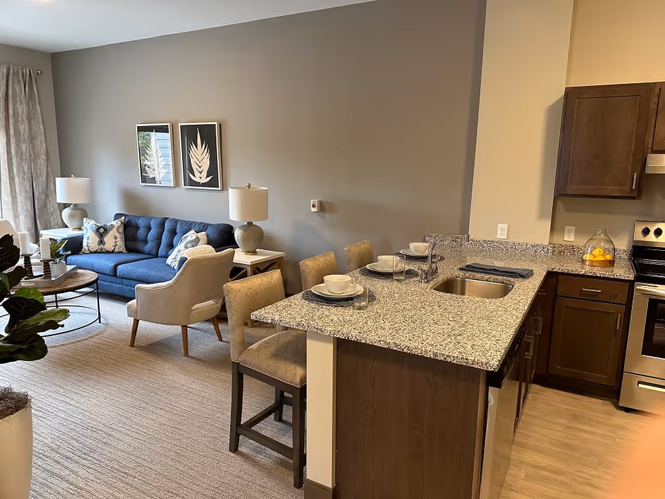 Open-concept living room and kitchen with a blue sofa, armchairs, wall art, and a granite-topped breakfast bar set with dishes.