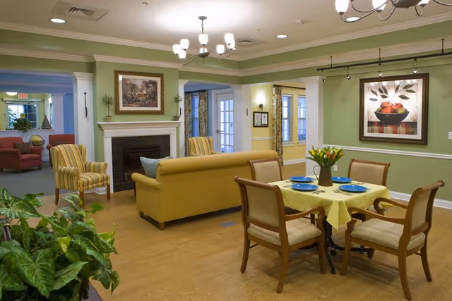 A cozy senior living common area with green walls and wooden flooring. The room features a yellow sofa, two striped armchairs, a small dining table set with four chairs and blue plates, a fireplace with a framed painting above it, and a large framed artwork on the wall. There are plants in the foreground and natural light coming through the windows with patterned curtains.