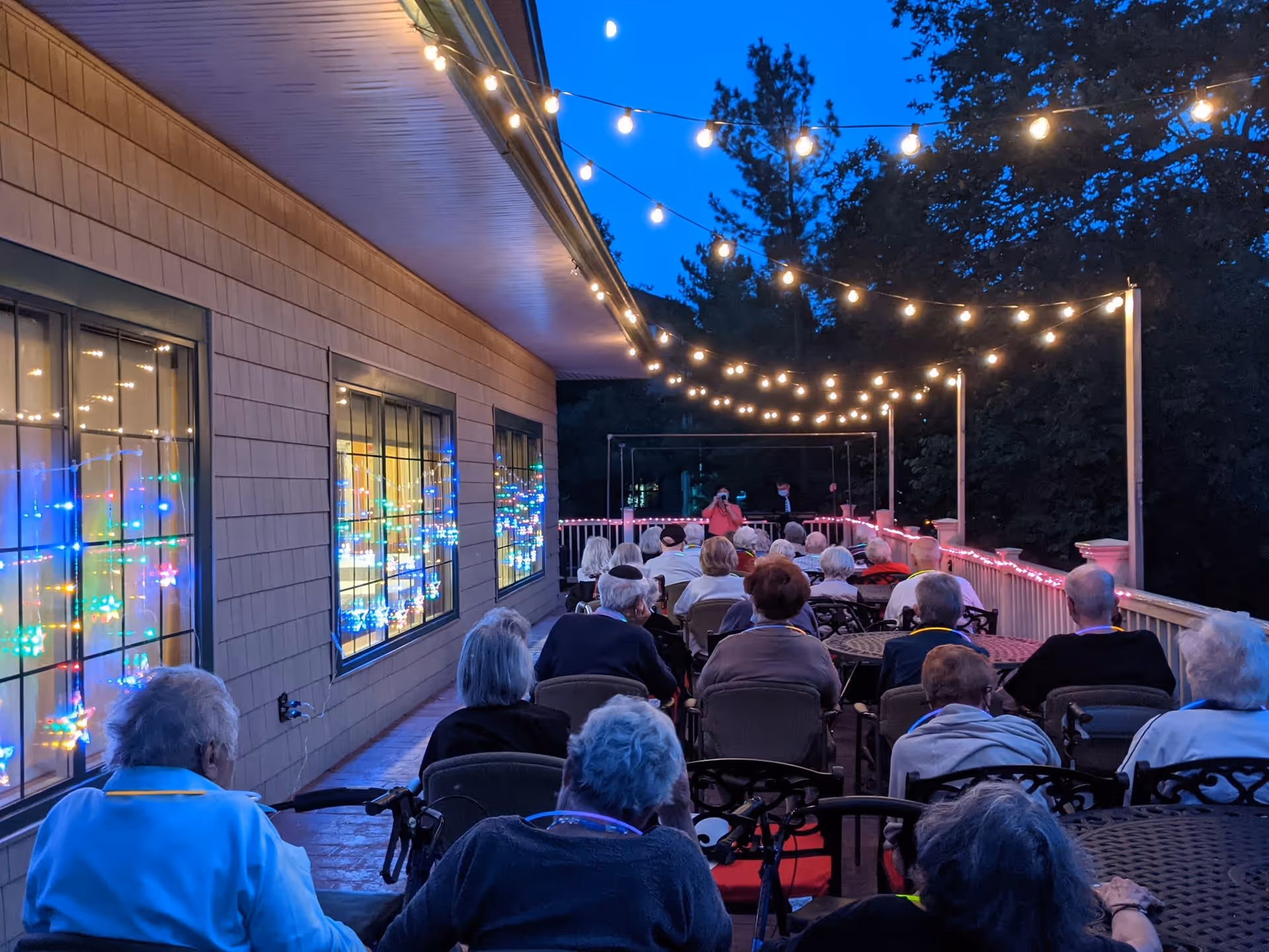 A group of elderly people seated outdoors on a patio at dusk, facing a person speaking or performing at the front. String lights are hung overhead, and colorful lights are visible through the windows on the left side of the building.