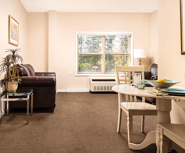 A cozy living room area with a brown leather sofa, a glass side table with a potted plant, a window with blinds letting in natural light, and a round white dining table with two chairs. The room has beige walls and carpeted flooring.