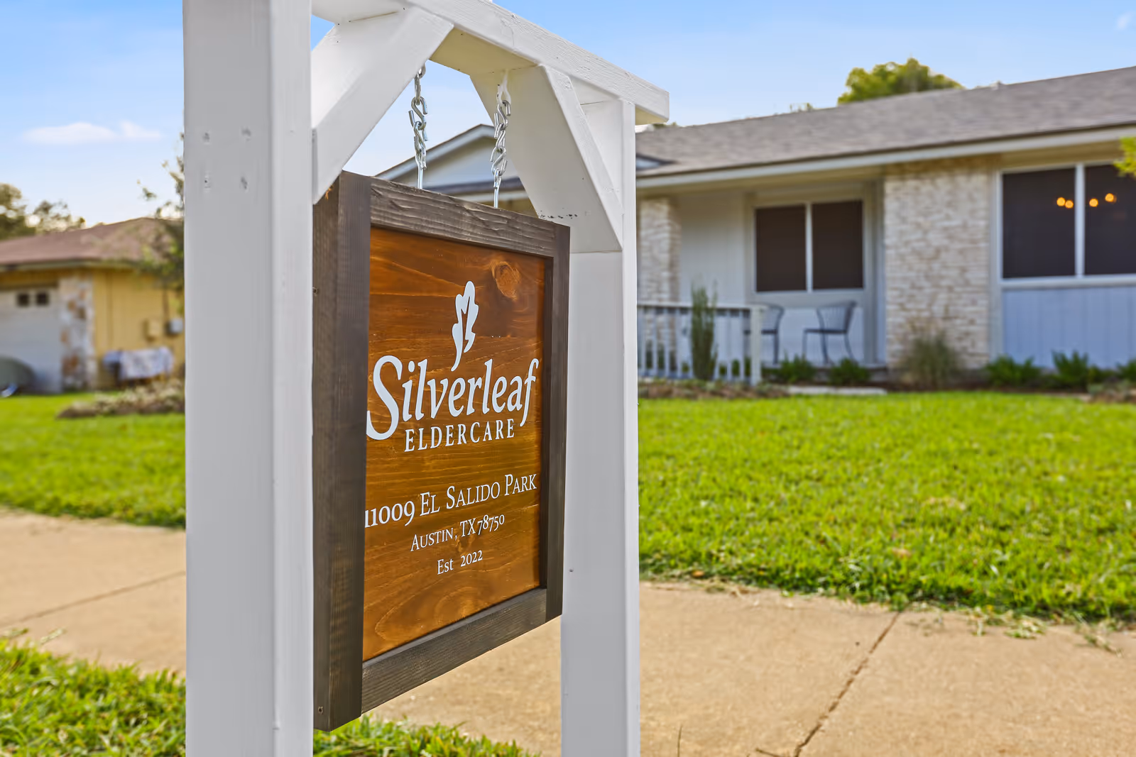 A wooden sign hanging from a white frame in front of a single-story building with a lawn and sidewalk. The sign reads 'Silverleaf Eldercare, 11009 El Salido Park, Austin, TX 78750, Est. 2022'.