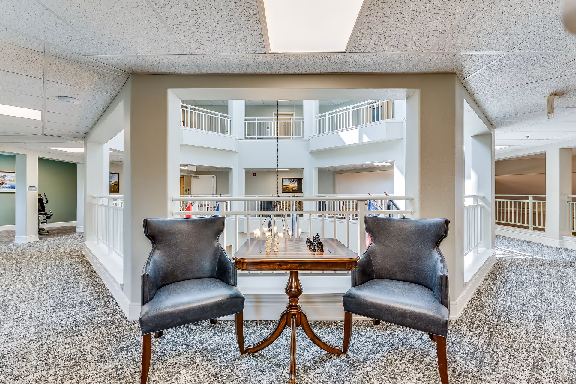 Two dark leather chairs facing each other with a wooden table in between, set up for a chess game in a bright, open indoor area with white railings and a view of multiple floors in the background.