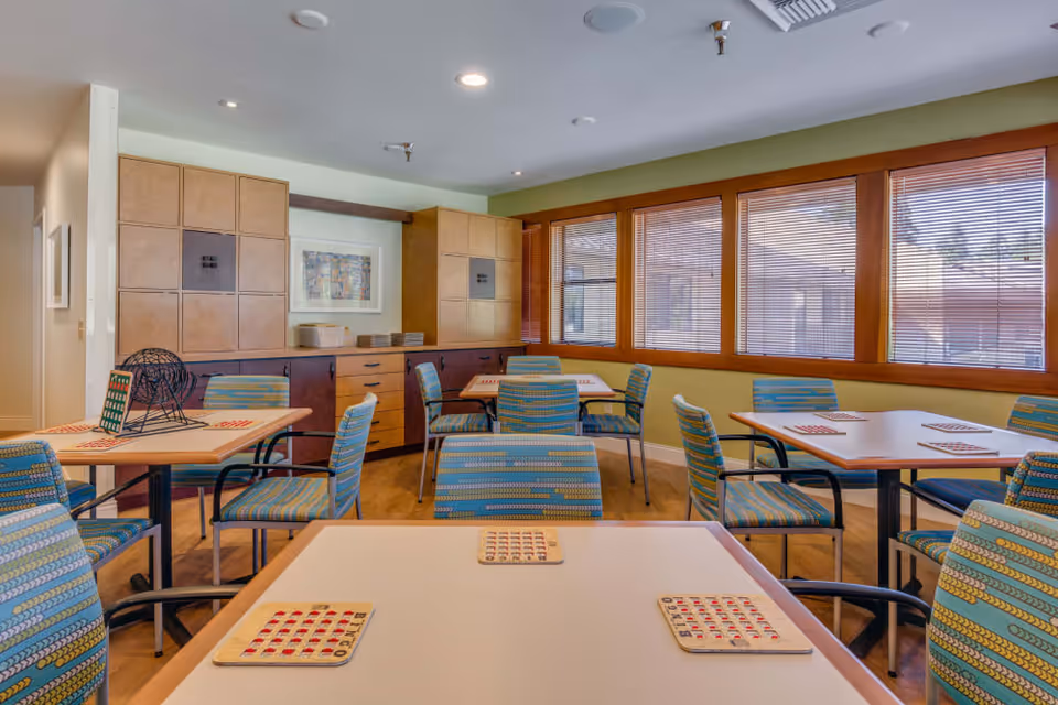 Well-lit community activity/dining room with several tables and striped upholstered chairs set up for bingo, large windows with blinds, and built-in cabinetry.