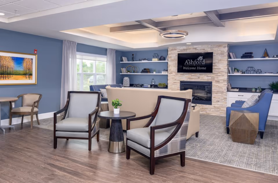 A bright, modern common lounge with armchairs, sofas, built-in shelving, a fireplace and a TV reading "The Ashford Welcome Home".