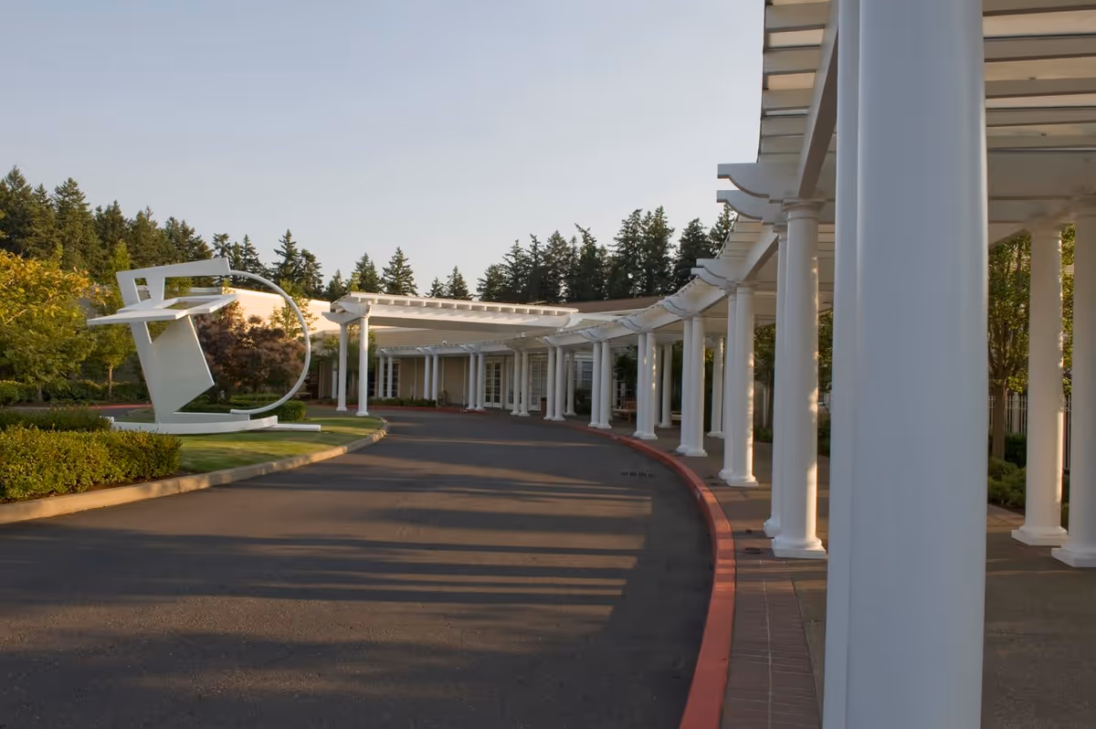 Curved driveway entrance to a building with white columns supporting a pergola-style roof, surrounded by greenery and trees, with a modern white sculpture on the left side.