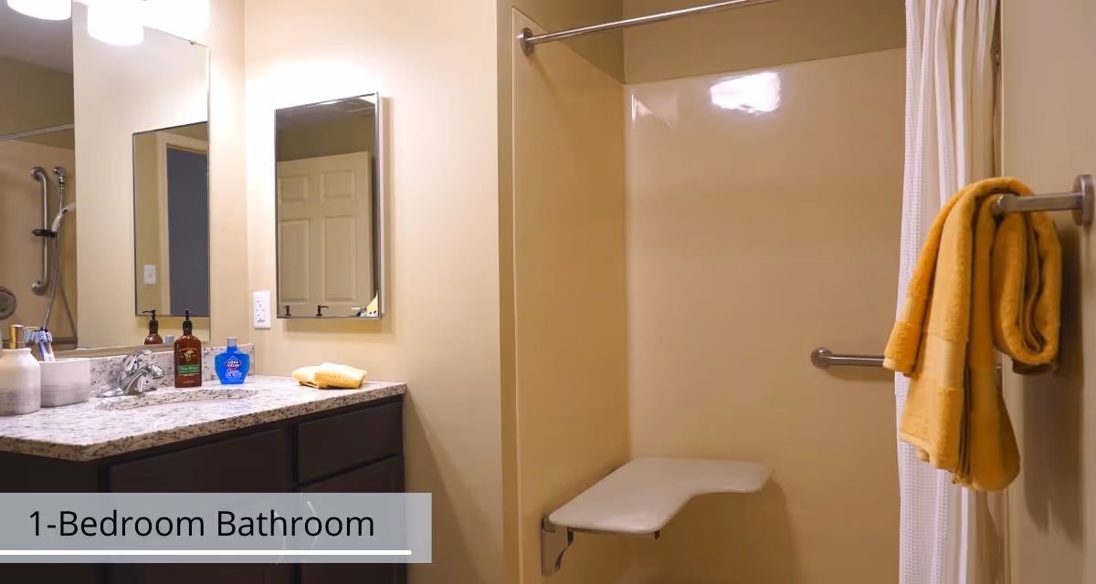 A bathroom with a granite countertop vanity featuring a sink, soap dispensers, and a small towel. There is a large mirror above the vanity and a smaller mirror on the adjacent wall. The shower area has a white fold-down seat, grab bars, and a white shower curtain. Yellow towels hang on a towel bar next to the shower.