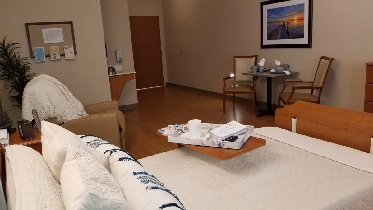 A cozy senior living bedroom featuring a bed with white bedding and pillows, a small overbed table with a cup, glasses, and a remote control. There is a comfortable armchair with a white throw blanket, a small round table with two chairs set with cups and napkins, a framed picture of a pier at sunset on the wall, and a wooden dresser. The room has wooden flooring and beige walls.