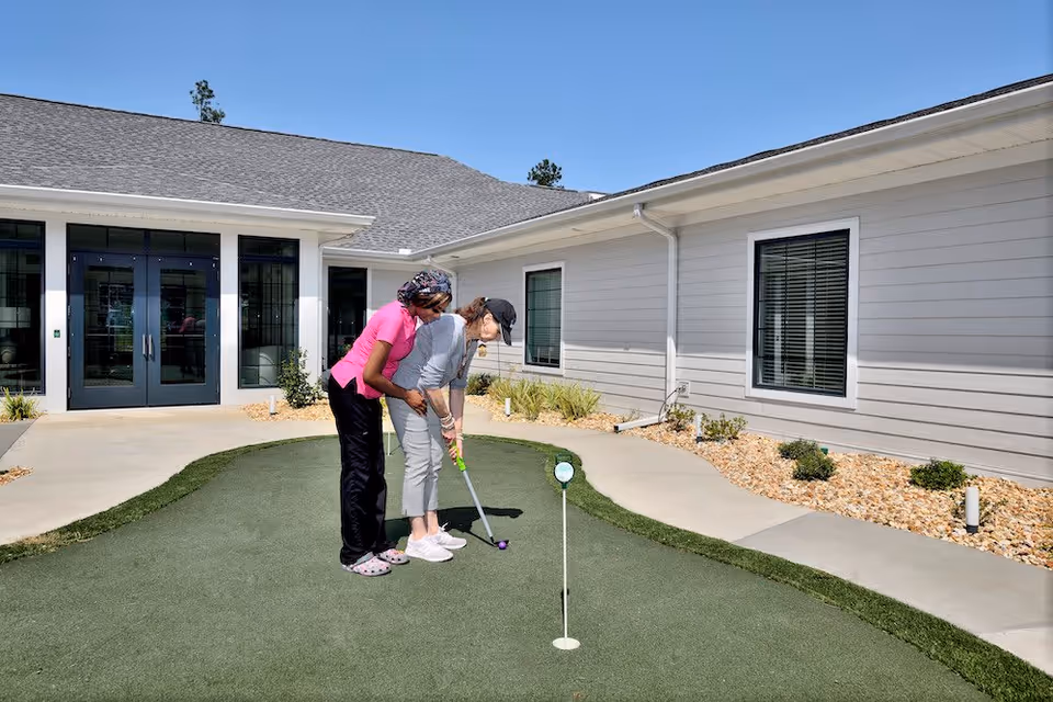 Two women playing mini golf on a putting green outside a single-story building with light gray siding and large windows under a clear blue sky. One woman is assisting the other with her golf stance.