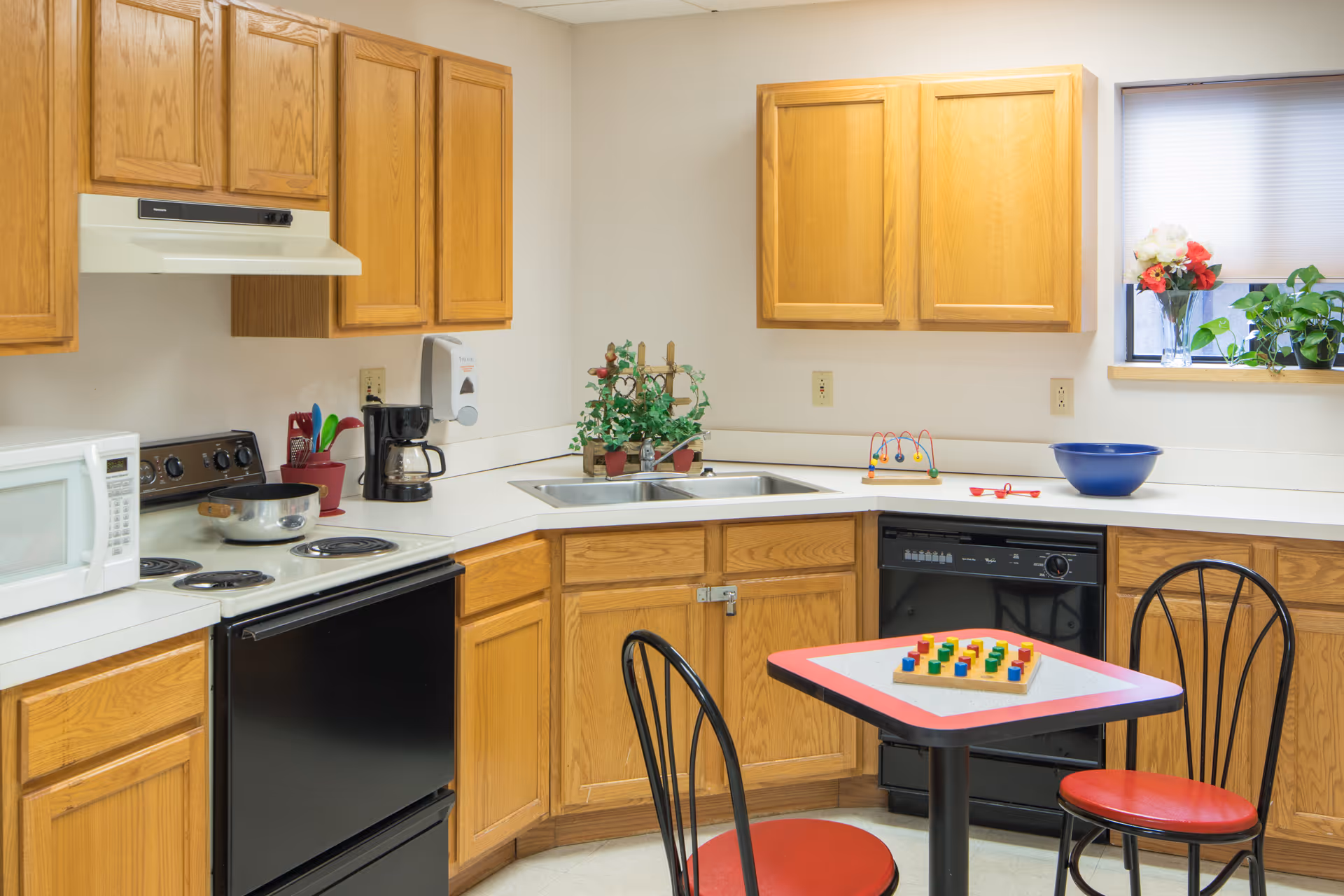 Kitchen with oak cabinets, stove, microwave, sink, dishwasher and a small table with two red chairs and a children's peg toy.
