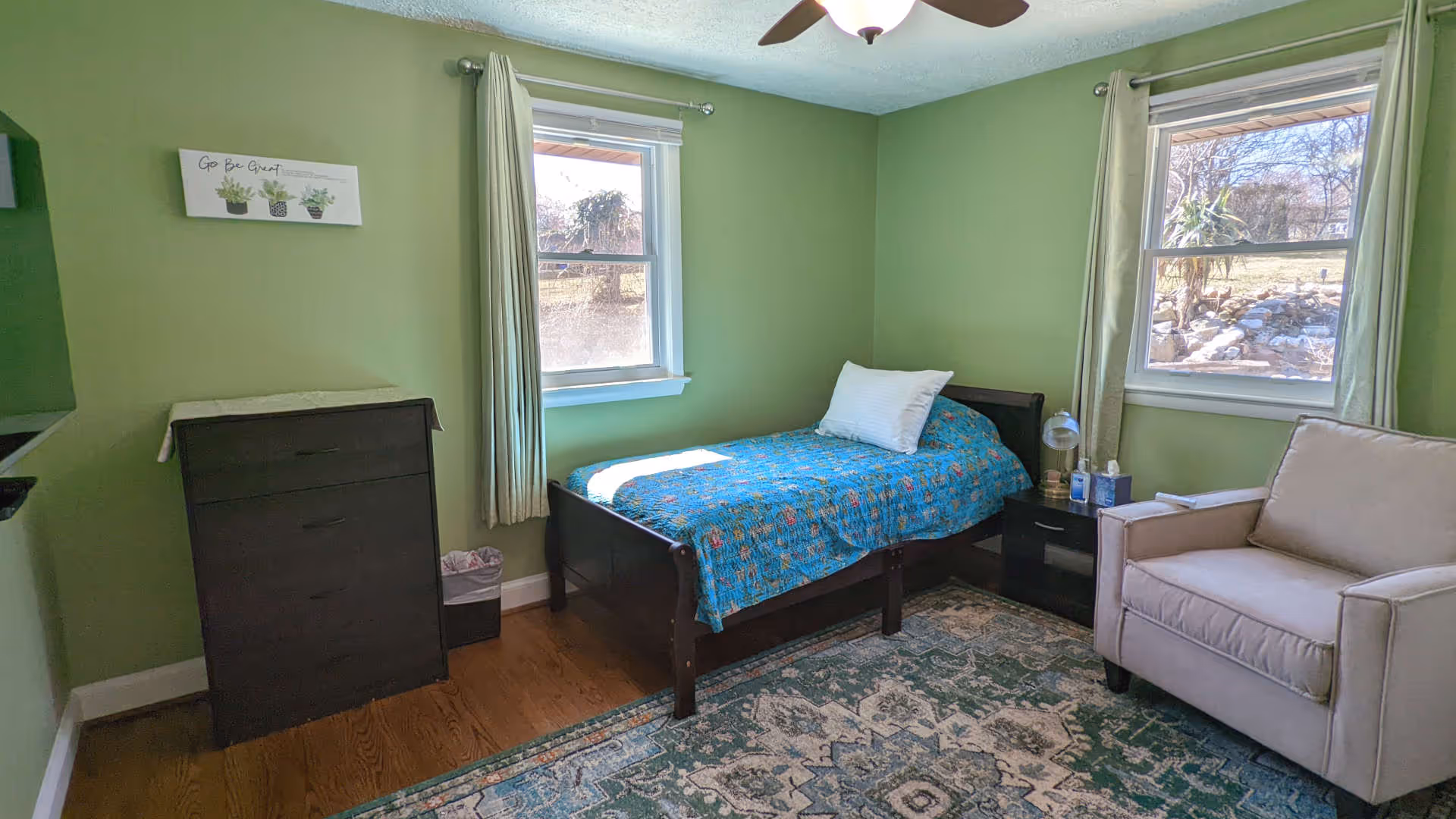 A small bedroom with green walls and two windows with light green curtains. The room contains a single bed with a blue floral bedspread and a white pillow, a beige armchair, a dark wooden dresser, and a matching nightstand with a lamp and some items on it. A patterned rug covers part of the wooden floor.