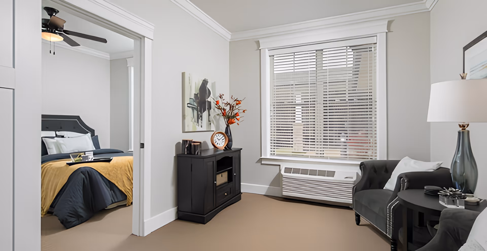 A cozy living area with a dark armchair, side table with a lamp, and a black cabinet with decorative items including a clock and a vase with orange flowers. A large window with blinds is visible, and through an open doorway, a bedroom with a bed covered in dark and mustard yellow bedding is seen.