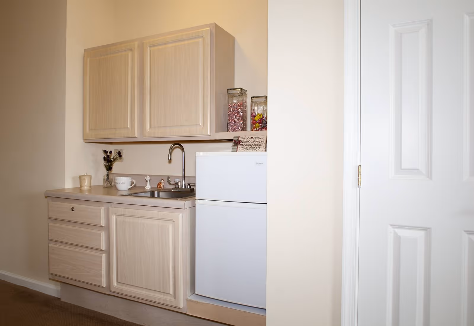 Small kitchenette area with light wood cabinets, a countertop with a sink and faucet, a small white refrigerator, and a few decorative items including a cup, a small vase with flowers, and two glass jars filled with candy.