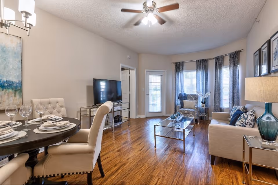 A bright and cozy living and dining area with wooden flooring. The dining area features a round table set with plates, napkins, and wine glasses, surrounded by upholstered chairs. The living area has a beige sofa with patterned cushions, a glass coffee table, an armchair, and a TV on a stand. Large windows with blue curtains allow natural light to fill the room, and a ceiling fan with lights is mounted on the ceiling.