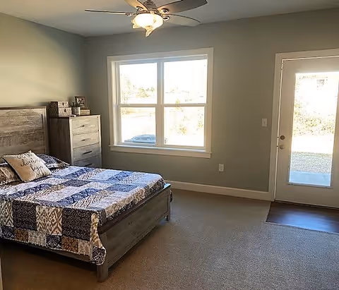 Sunny bedroom with a bed, dresser, ceiling fan, a large window, and a glass door leading outside.