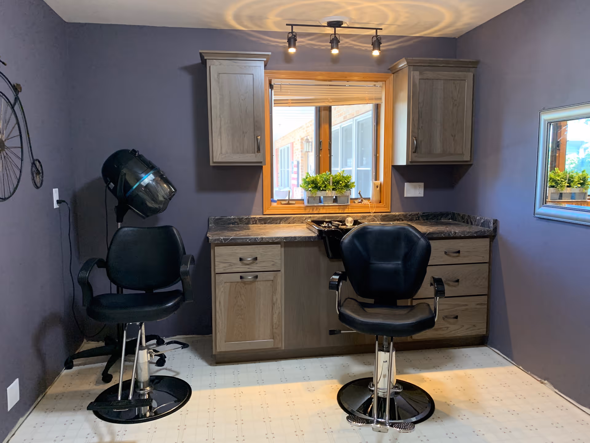 A small salon area with two black salon chairs, a hair dryer, wooden cabinets, a countertop, and a window with potted plants on the sill. The walls are painted purple and there is a mirror on the right wall.