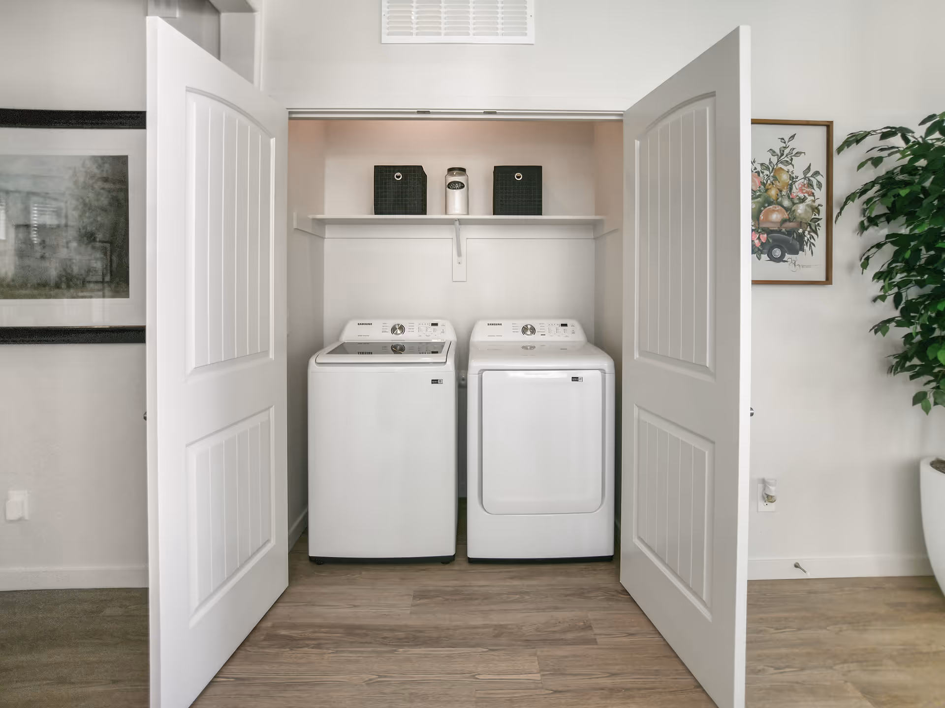 Open closet-style laundry area with a washer and dryer side by side under a shelf holding storage baskets, framed art and a plant visible nearby.