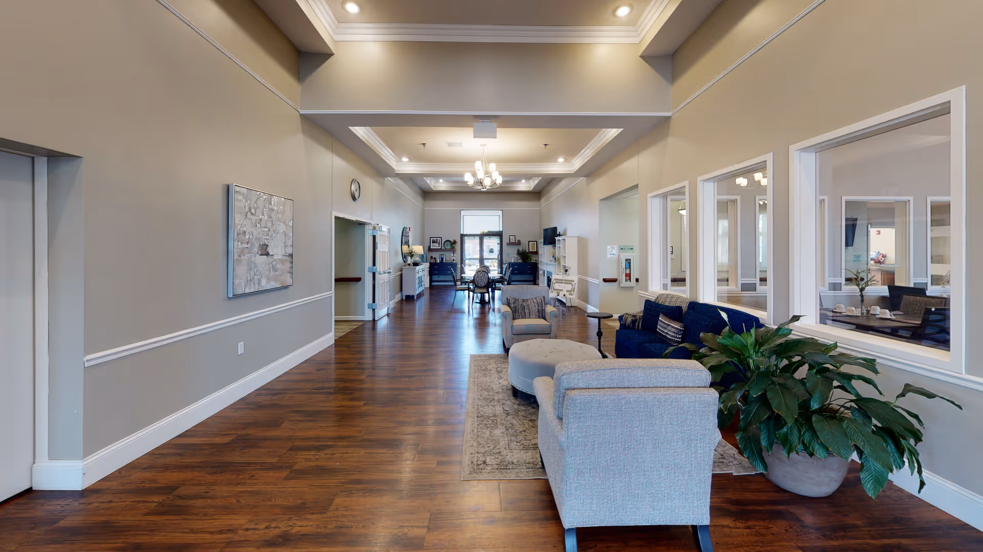 A spacious senior living facility common area with wooden flooring, beige walls, and recessed lighting. The room features comfortable seating including armchairs, a sofa, and an ottoman arranged on a rug. Large windows on the right wall look into adjacent rooms with tables and chairs. There are plants and decorative artwork on the walls, and a chandelier hangs from the ceiling. The far end of the room has more seating and a door leading outside.