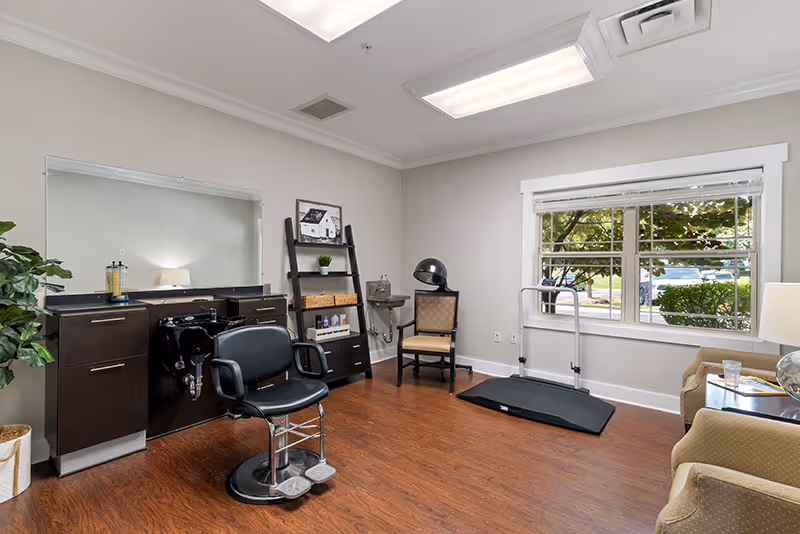 A bright interior salon room with a styling chair and shampoo station, hood dryer, shelving, seating and a window overlooking trees.