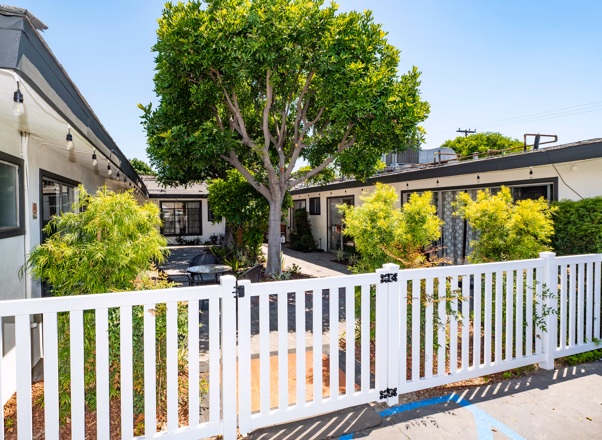 Outdoor courtyard area of a senior living facility with a white picket fence in the foreground, green trees and bushes, and a paved walkway surrounded by single-story buildings under a clear blue sky.