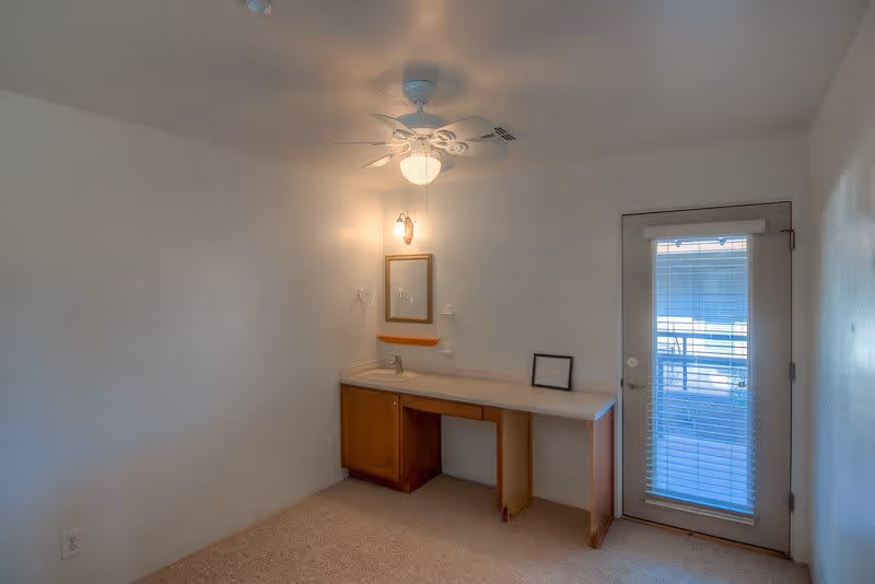 A small room with beige carpet, white walls, and a ceiling fan with a light. There is a wooden vanity with a sink, a small mirror above it, and a light fixture on the wall. To the right, there is a door with a window covered by blinds, leading outside.