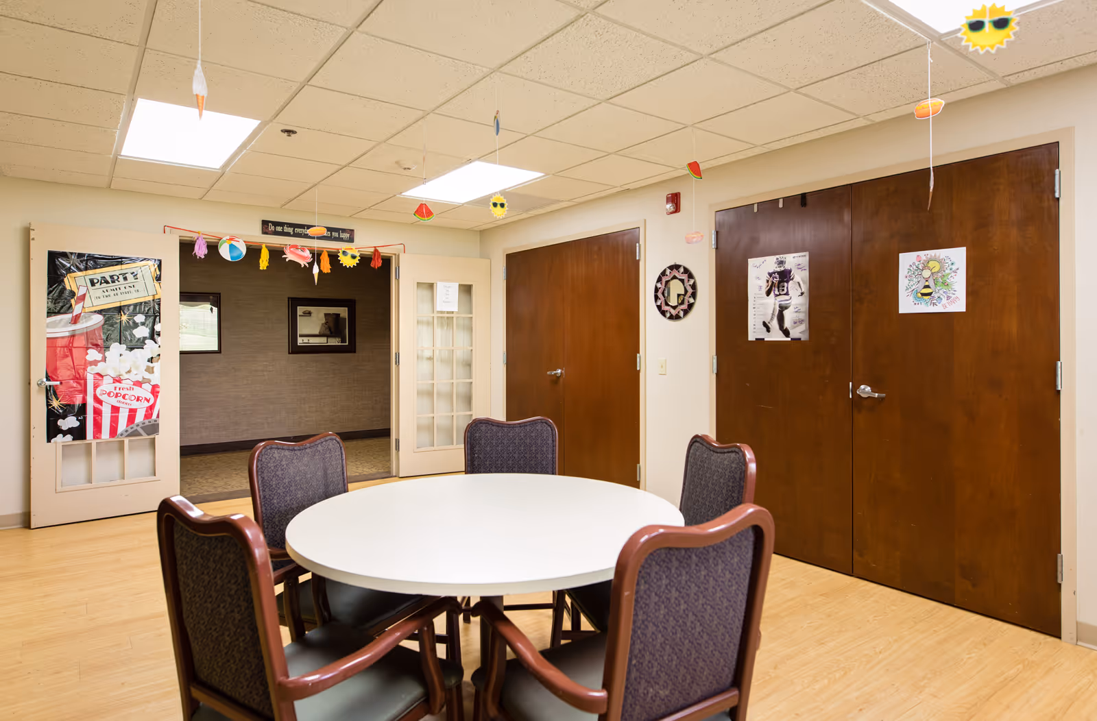 A room with a round white table surrounded by five chairs with purple upholstery and wooden frames. The room has light-colored wooden flooring and a drop ceiling with fluorescent lights. There are two closed wooden double doors on one wall, each decorated with posters. Another wall has glass-paneled double doors, one decorated with a popcorn-themed poster. Colorful hanging decorations shaped like summer items such as a sun with sunglasses, watermelon slices, and ice cream cones hang from the ceiling. A small decorative mirror is mounted on the wall between the wooden doors.