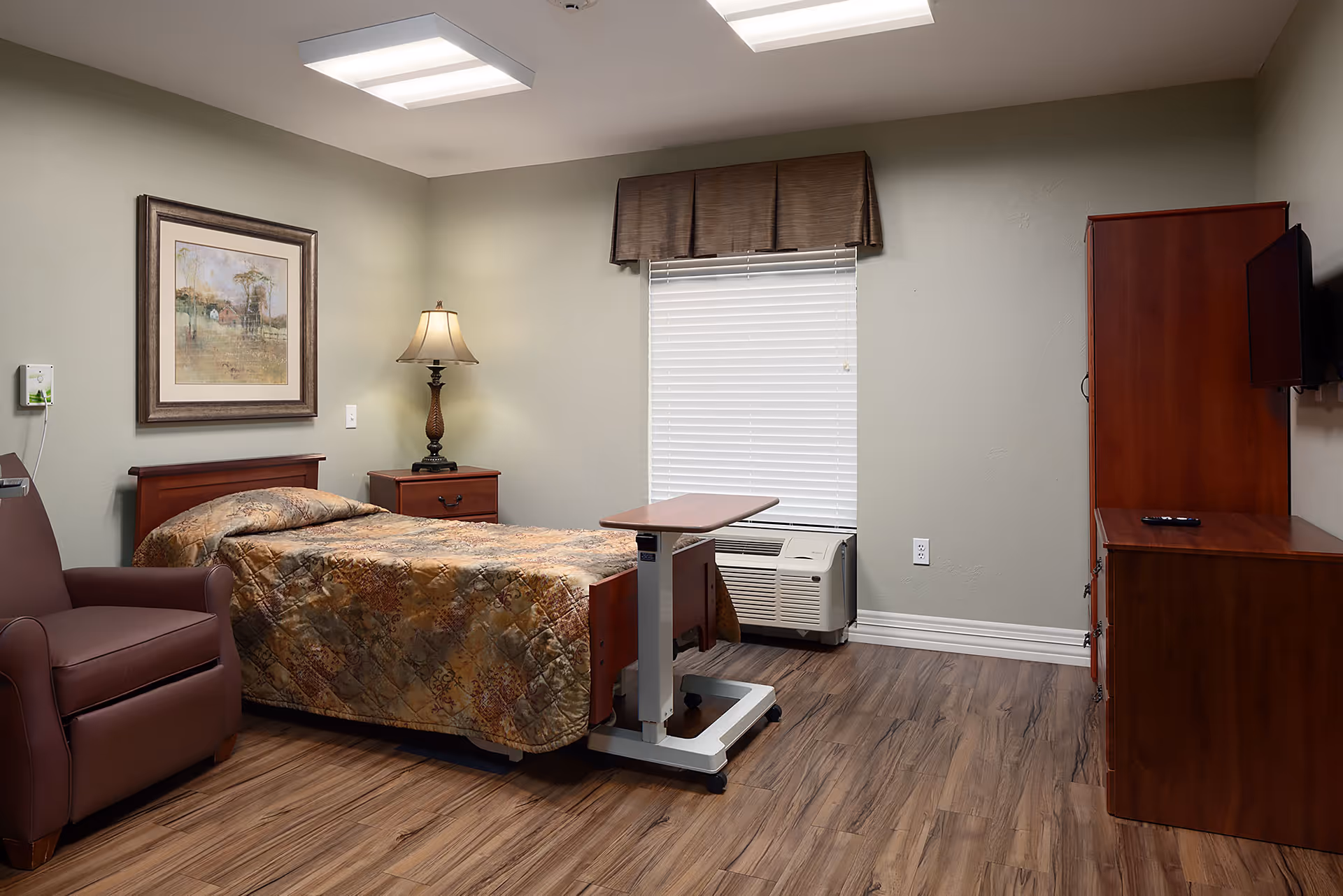 A furnished private bedroom in a senior facility with a single bed, overbed table, recliner, nightstand with lamp, dresser-mounted TV, and a window.