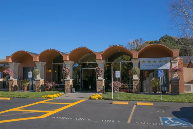 Front entrance of a post-acute care facility with an arched covered walkway, potted plants, and marked ambulance parking spaces.