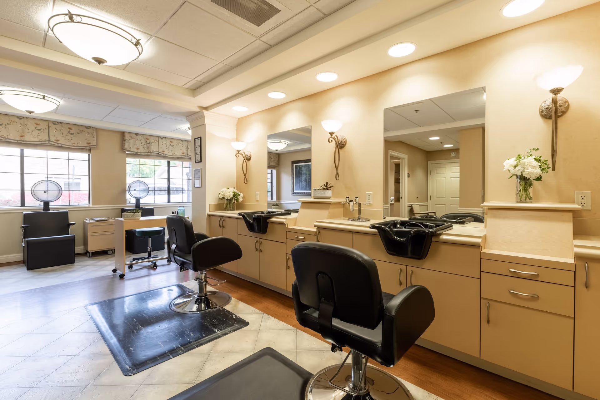 Interior view of a senior living facility's hair salon area with black salon chairs, sinks for hair washing, large mirrors, and windows letting in natural light. The room is well-lit with ceiling lights and wall sconces, and there are flowers placed on the counters.