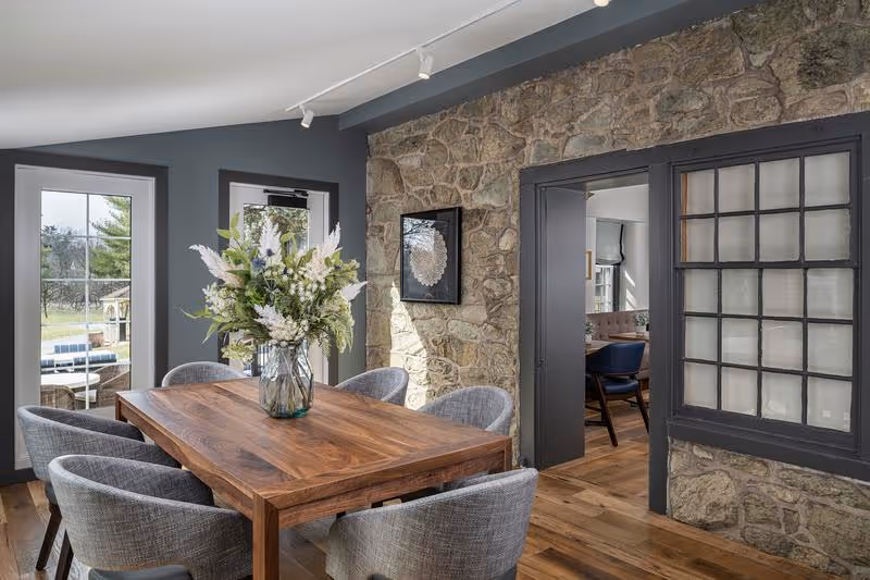 Dining room with a wooden table surrounded by upholstered chairs, a vase of flowers, a stone accent wall, and doors leading outside and to another room.