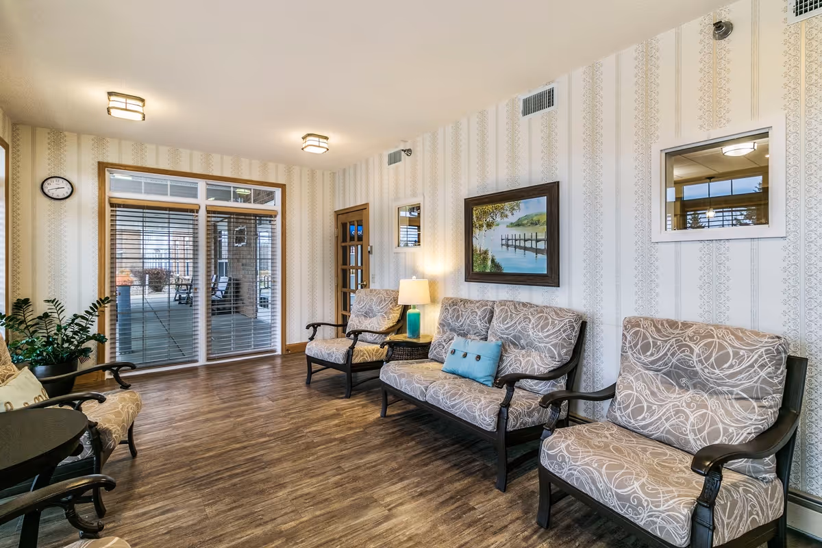 A cozy sitting area in St. Andrew’s Village featuring patterned cushioned chairs and a loveseat with dark wooden frames, a small round table, a potted plant, a table lamp with a blue base, and a framed painting of a lakeside scene on the wallpapered wall. The room has wood flooring, two ceiling lights, a wall clock, and large windows with blinds looking out to an outdoor covered patio area.