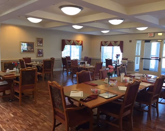 A dining room in a senior living facility with multiple wooden tables and chairs arranged neatly. Each table is set with placemats, napkins, menus, and small flower vases. The room has large windows with decorative valances, framed artwork on the walls, and ceiling lights providing bright illumination. There is a glass door leading outside or to another area.