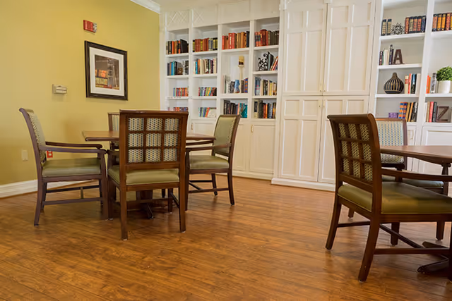 Interior room with wooden floor, several wooden chairs with green cushions around wooden tables, and white built-in bookshelves filled with books and decorative items against a yellow wall with framed artwork.