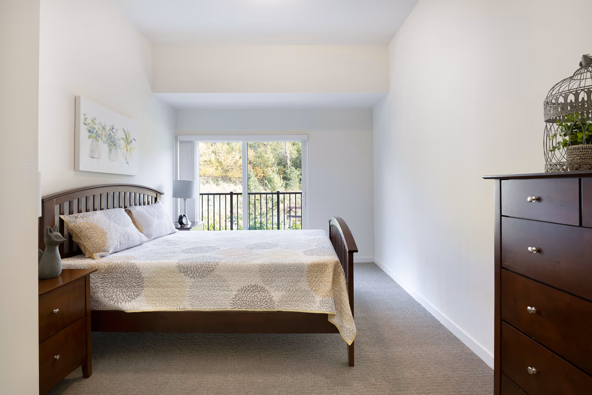 A bright and clean bedroom with a wooden bed frame and a bed covered with a white and gray patterned quilt. There are two pillows on the bed. To the left of the bed is a wooden nightstand with a small decorative cat figurine and a lamp. On the right side of the room is a tall wooden dresser with a decorative birdcage containing a green plant on top. The room has a large window with vertical blinds that opens to a view of trees outside.