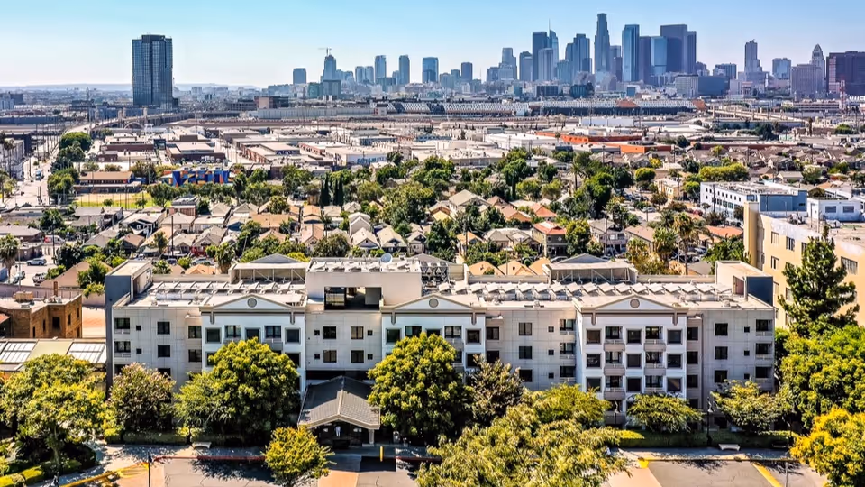 Aerial view of Sakura Intermediate Care Facility, a multi-story building surrounded by trees and greenery, with a city skyline in the background under a clear sky.