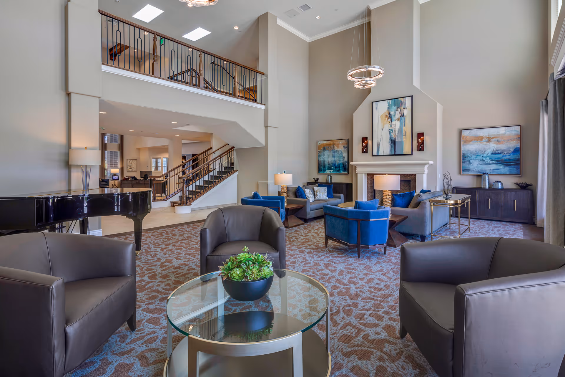 Spacious two-story communal living room with multiple seating areas, a fireplace, a grand piano, and an open staircase.