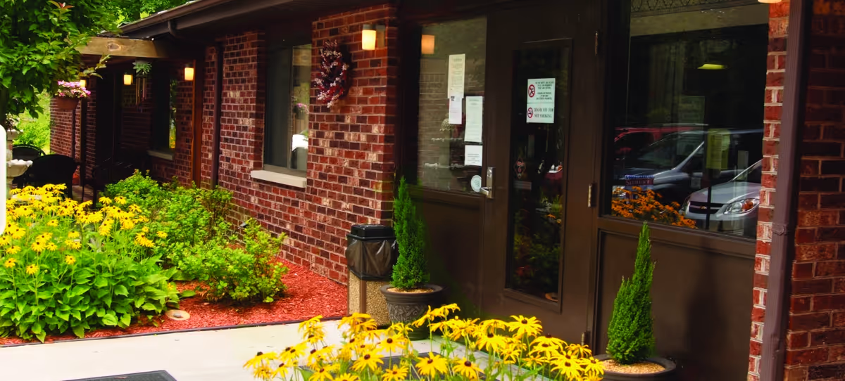 Brick exterior entrance with glass double doors, potted evergreens and beds of yellow flowers.