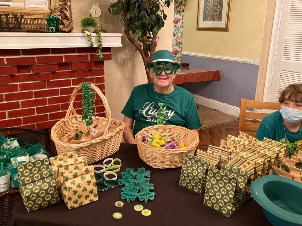 Two people sitting at a table decorated with St. Patrick's Day themed items including shamrock-shaped glasses, green hats, and green necklaces. The table is covered with various green and shamrock-patterned gift bags, bracelets, and small baskets filled with treats. The background shows a red brick fireplace and a room with light-colored walls and framed pictures.