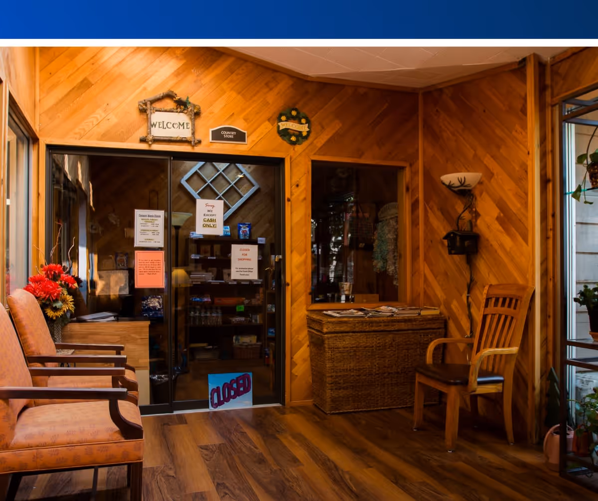 A cozy waiting area with wooden paneled walls and wood flooring. There are two upholstered chairs on the left and a wooden chair on the right. A wicker chest with magazines on top is against the wall. A glass door with signs indicating the store is closed and cash only leads to a small country store. Decorative welcome signs hang on the wall above the door.
